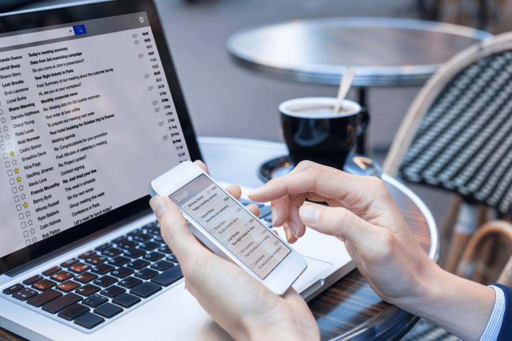 Laptop mit offenen E-Mails und Person mit Smartphone in der Hand, die E-Mail-Betreffzeilen liest