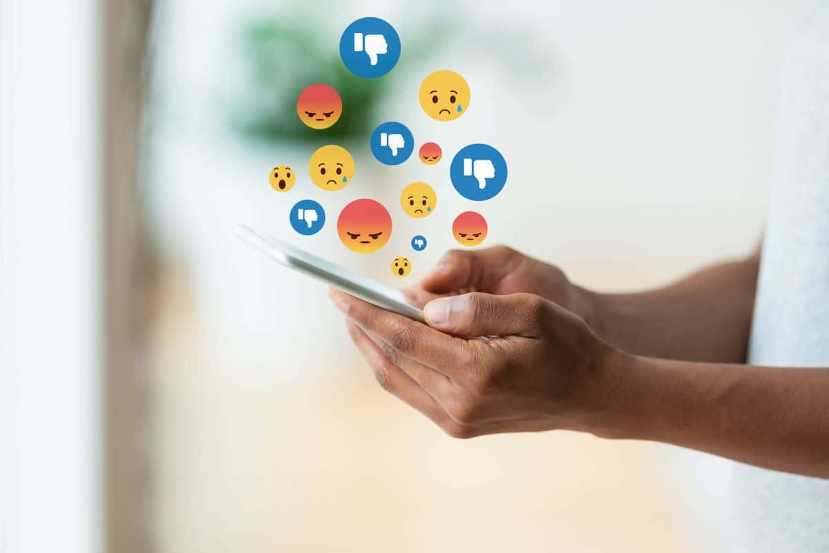Hände halte Smartphone, von dem aus Emojis als Zeichen für negative Kommentare aufsteigen