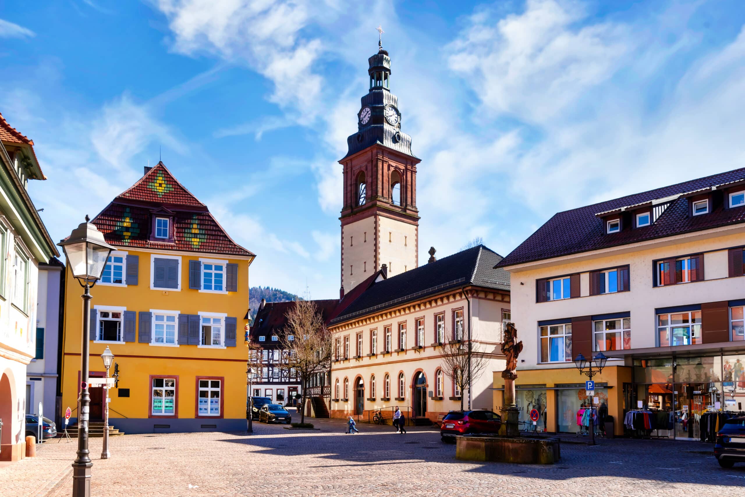 Auf der linken Seite ein dreistöckiges gelbes Haus, und neben an eine Kirche mit großem Kirchturm. Davor ein Marktplatz mit einem alten Brunnen.